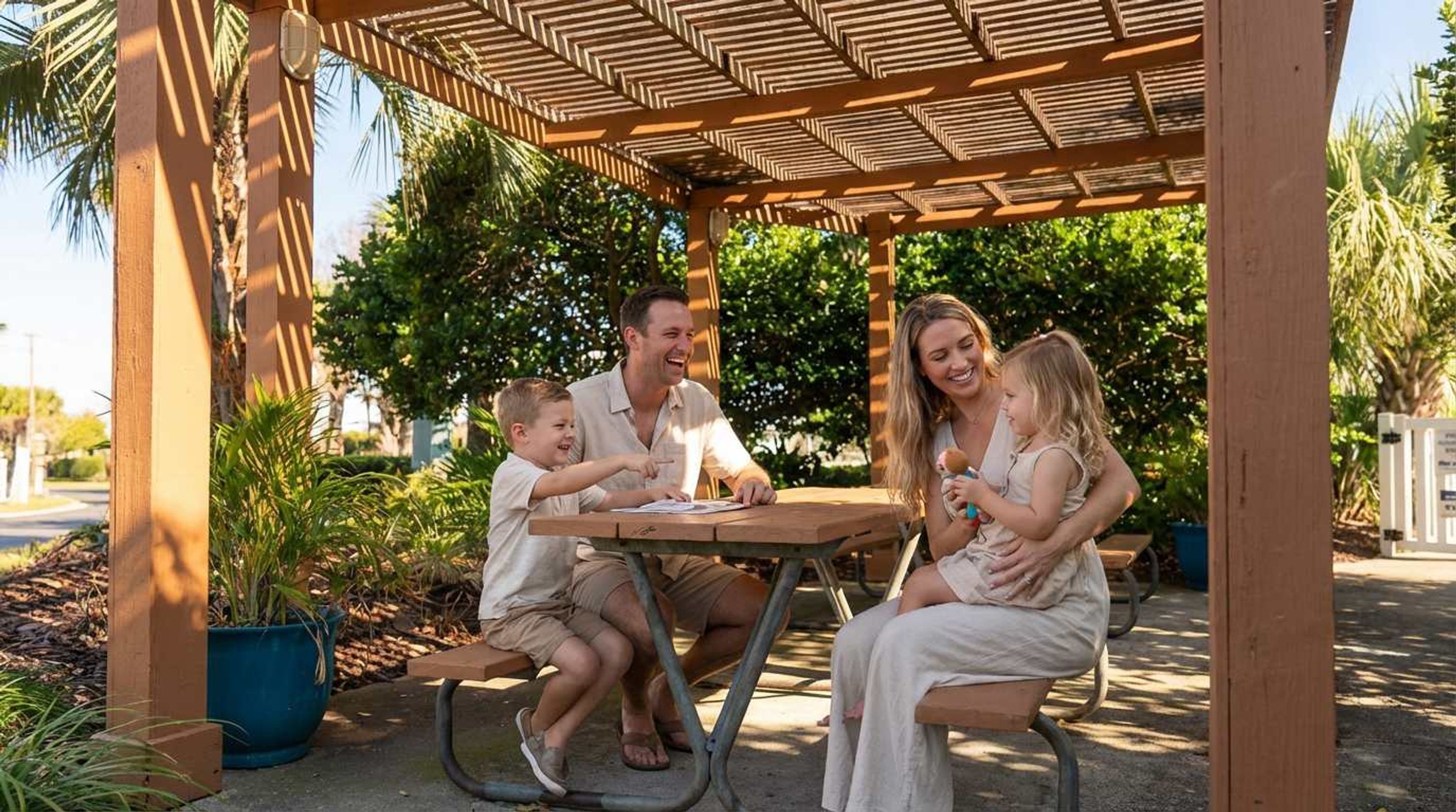 A family of four seated at a wooden table under a pergola with a view of lush greenery.