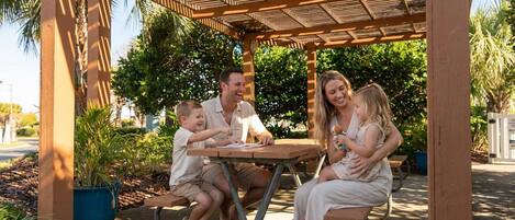 A family of four seated at a wooden table under a pergola with a view of lush greenery.