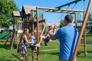 A playground with a wooden climbing structure and swings. - Steps to the Beach Pool Courts Family Fun (Destin)