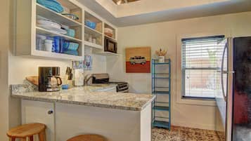 Cocina con encimera de granito, armarios blancos y microondas. Hay dos taburetes de madera y una ventana con persianas.