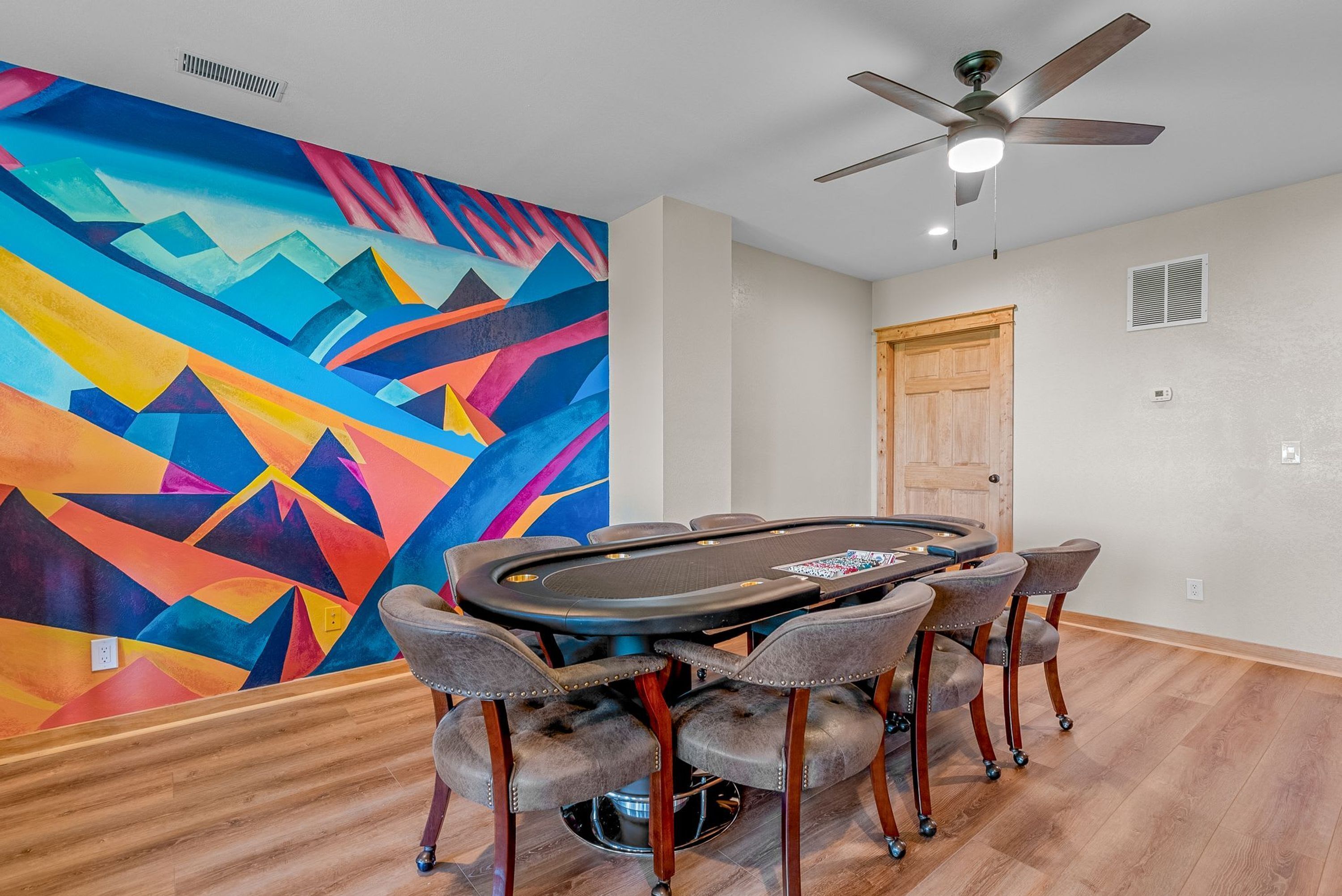 A room with a poker table, six chairs, a ceiling fan, and a colorful abstract mural on the wall.