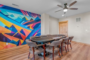 A room with a poker table, six chairs, a ceiling fan, and a colorful abstract mural on the wall.