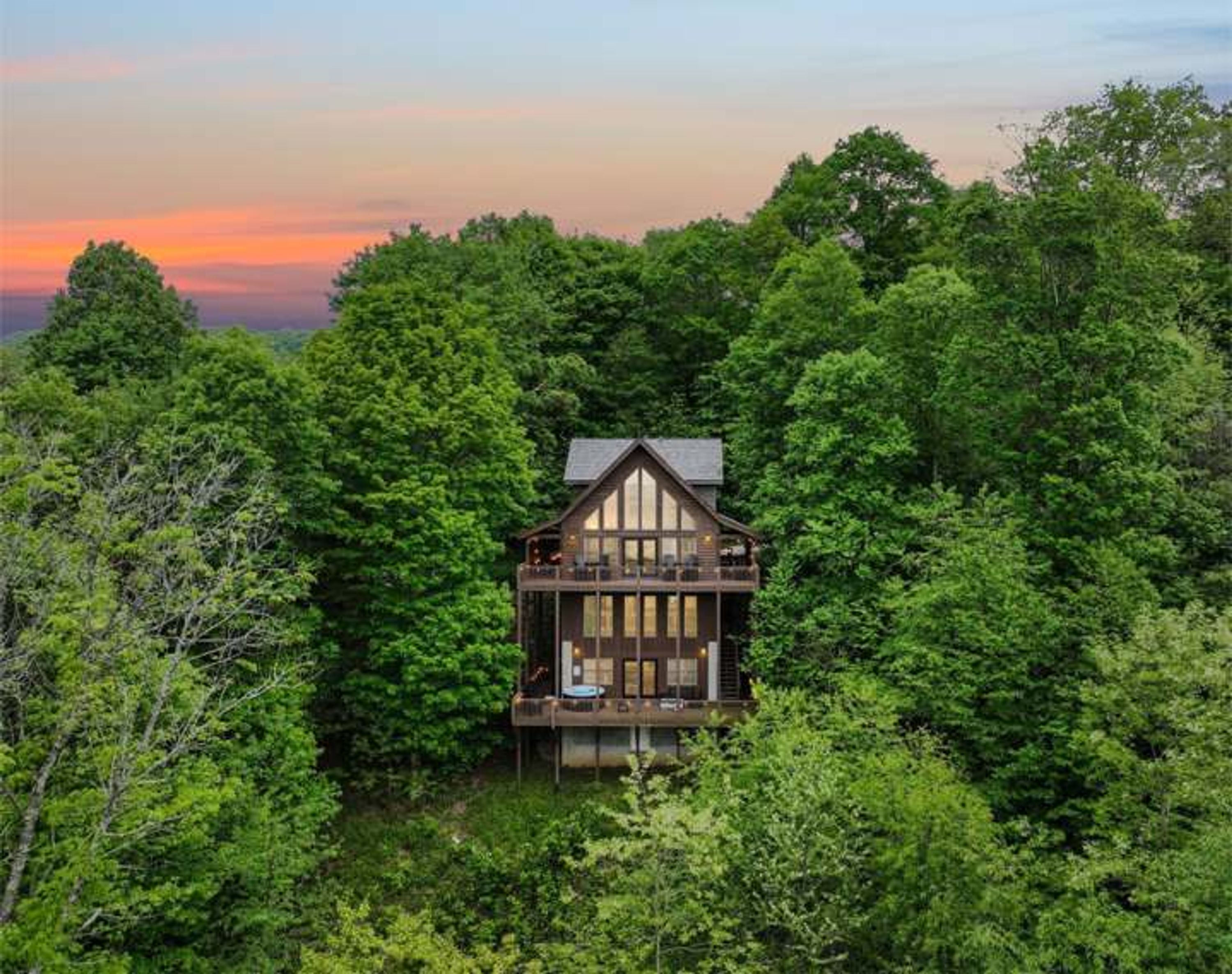 A cabin surrounded by dense forest, with a prominent front porch and a steep roof.