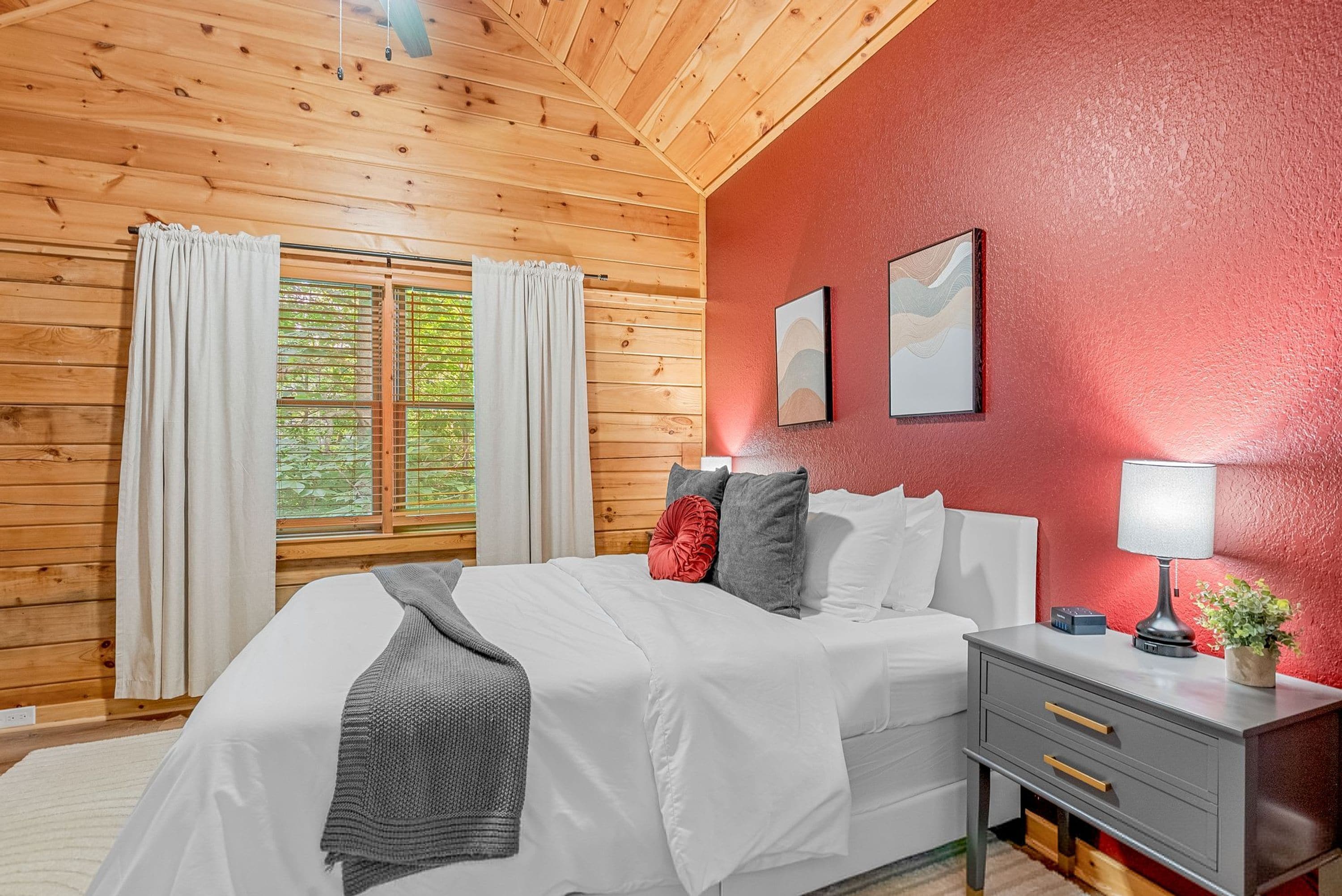 Un dormitorio con techo de madera, una cama con ropa blanca, una mesita de noche con lámpara y una pared roja.