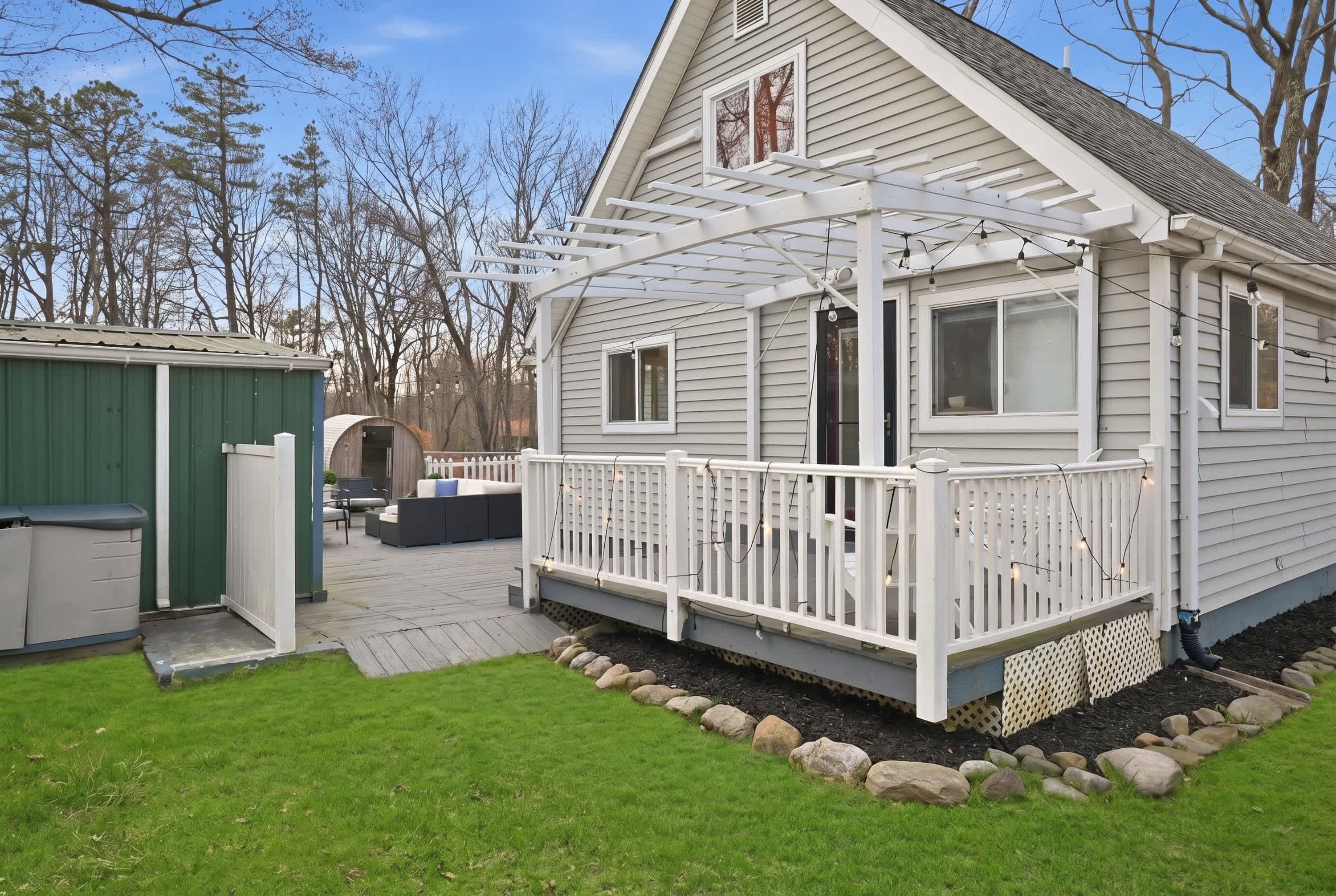 Une maison de plain-pied dotée d’un porche couvert, d’un petit abri de jardin et d’un espace patio aménagé avec du mobilier d’extérieur.