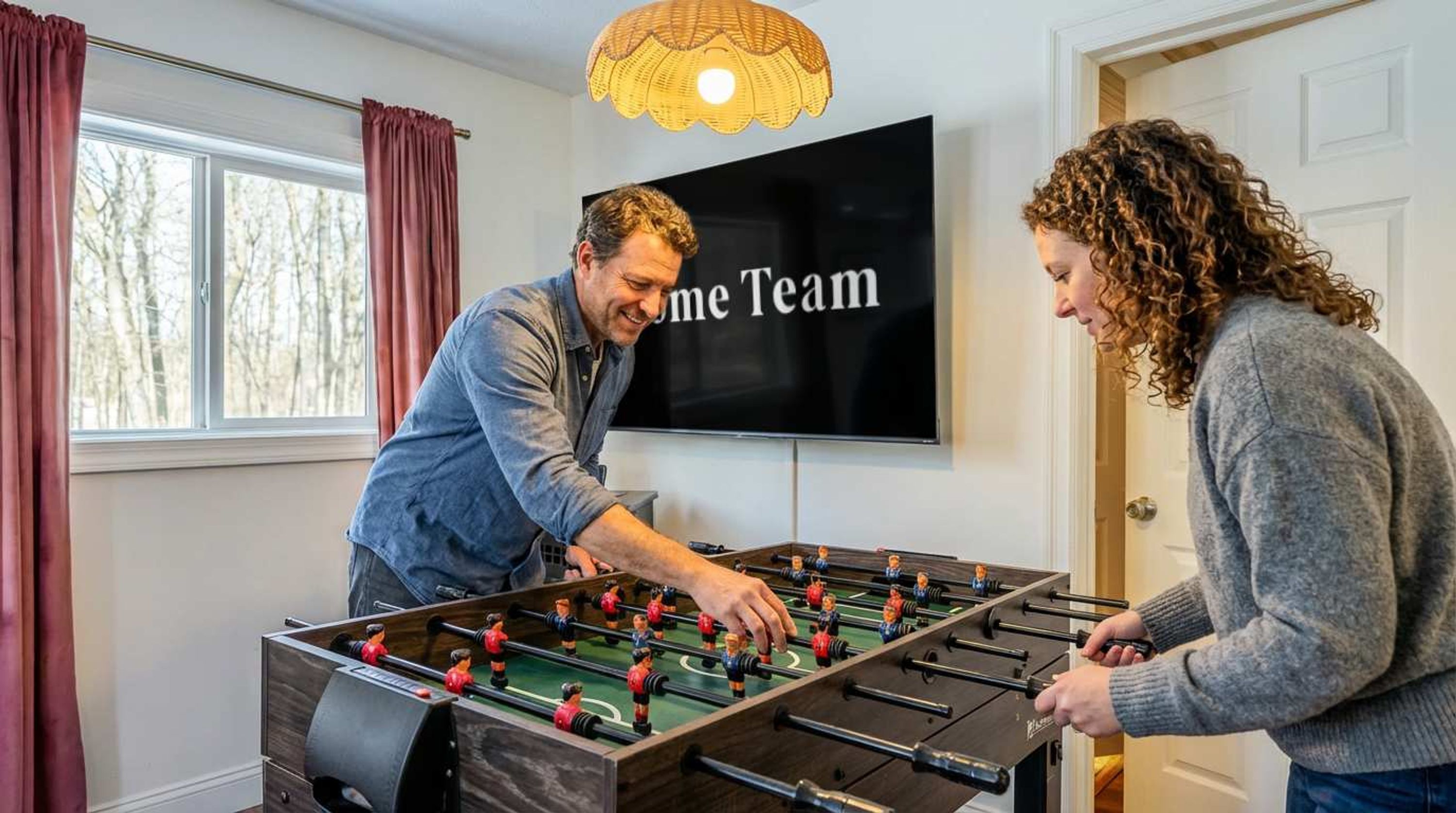 A room with a foosball table, a television displaying 'Welcome Team', and a window with curtains.