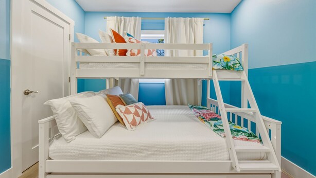 A bunk bed room with a white frame, a lower bed with white bedding and colorful pillows, and a bunk above with a floral bedspread. - Sports Court Playground Putt Putt Spa Pool (Bradenton)