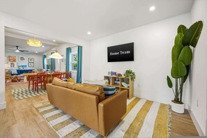 Una sala de estar moderna con un sofá, un comedor con sillas, una planta grande en maceta y un cartel que dice 'Home Team'.