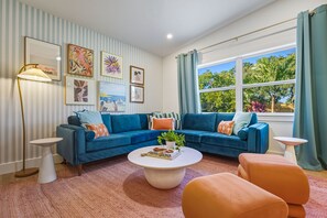 A living room with a blue sofa, a coffee table, and a large window with a view of trees. - Sports Court Playground Putt Putt Spa Pool (Bradenton)