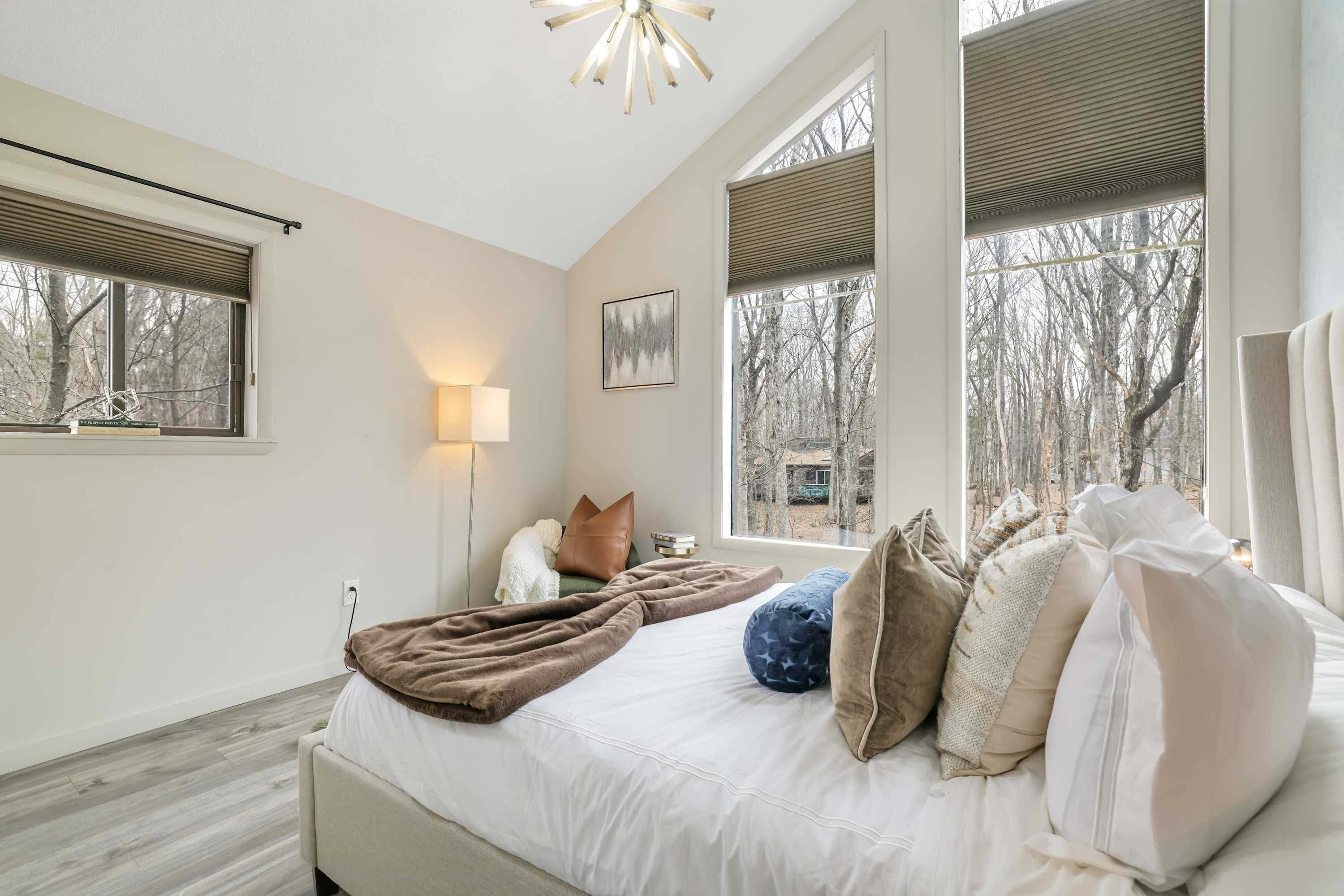 A bedroom with a large bed, multiple pillows, a bedside lamp, and a view of trees through the windows.