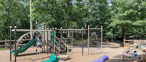 A playground with various play structures, including a slide and climbing frames, surrounded by trees.