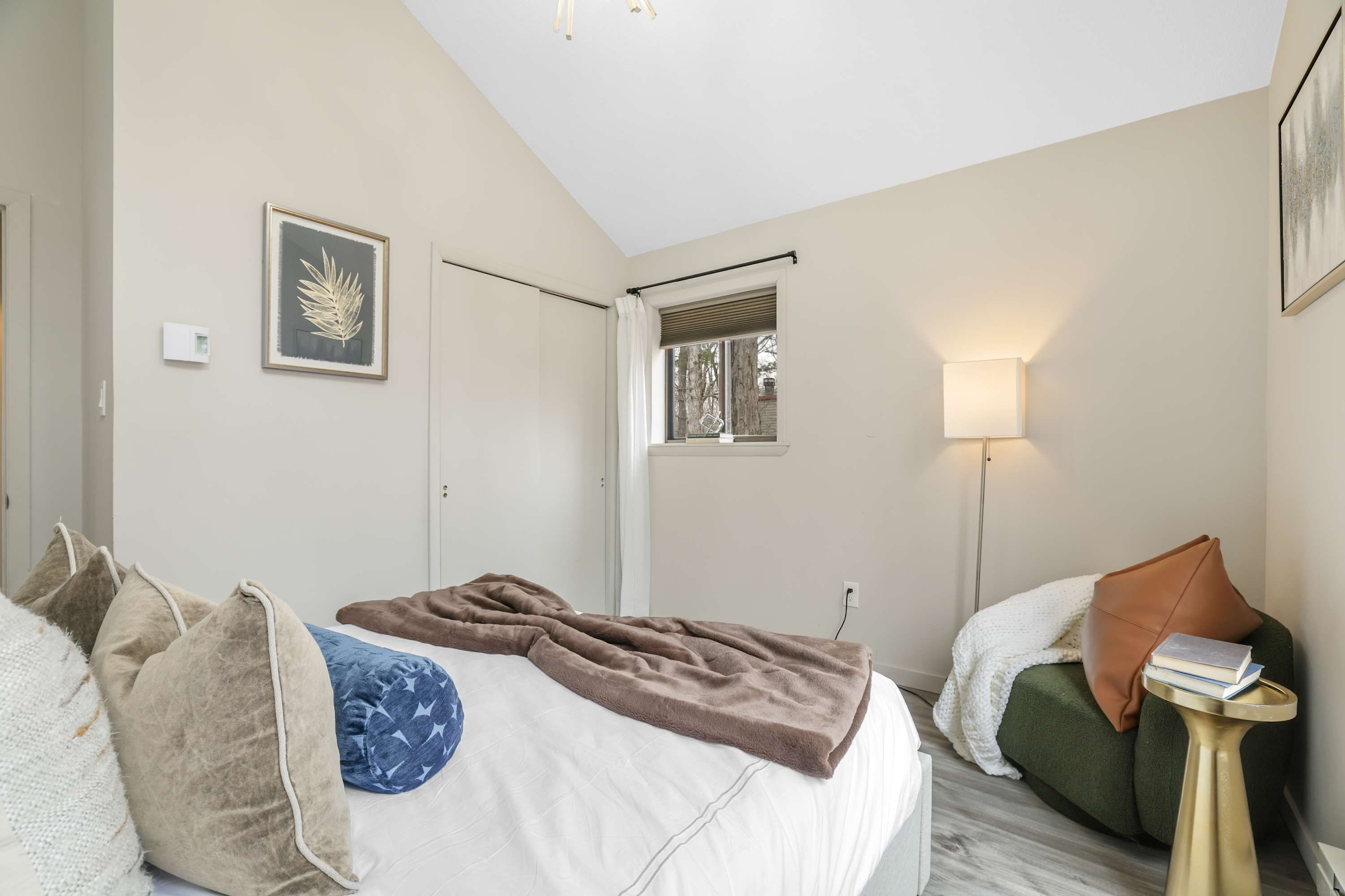 A bedroom with a bed, pillows, a blanket, a bedside lamp, a window, and a framed picture on the wall.
