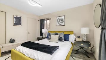 Habitación de hotel con cama, mesitas de noche, escritorio, silla, ventana con cortinas y obras de arte en las paredes.