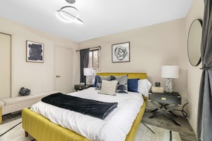Quarto de hotel com cama, mesas de cabeceira, escrivaninha, cadeira, janela com cortinas e obras de arte nas paredes.