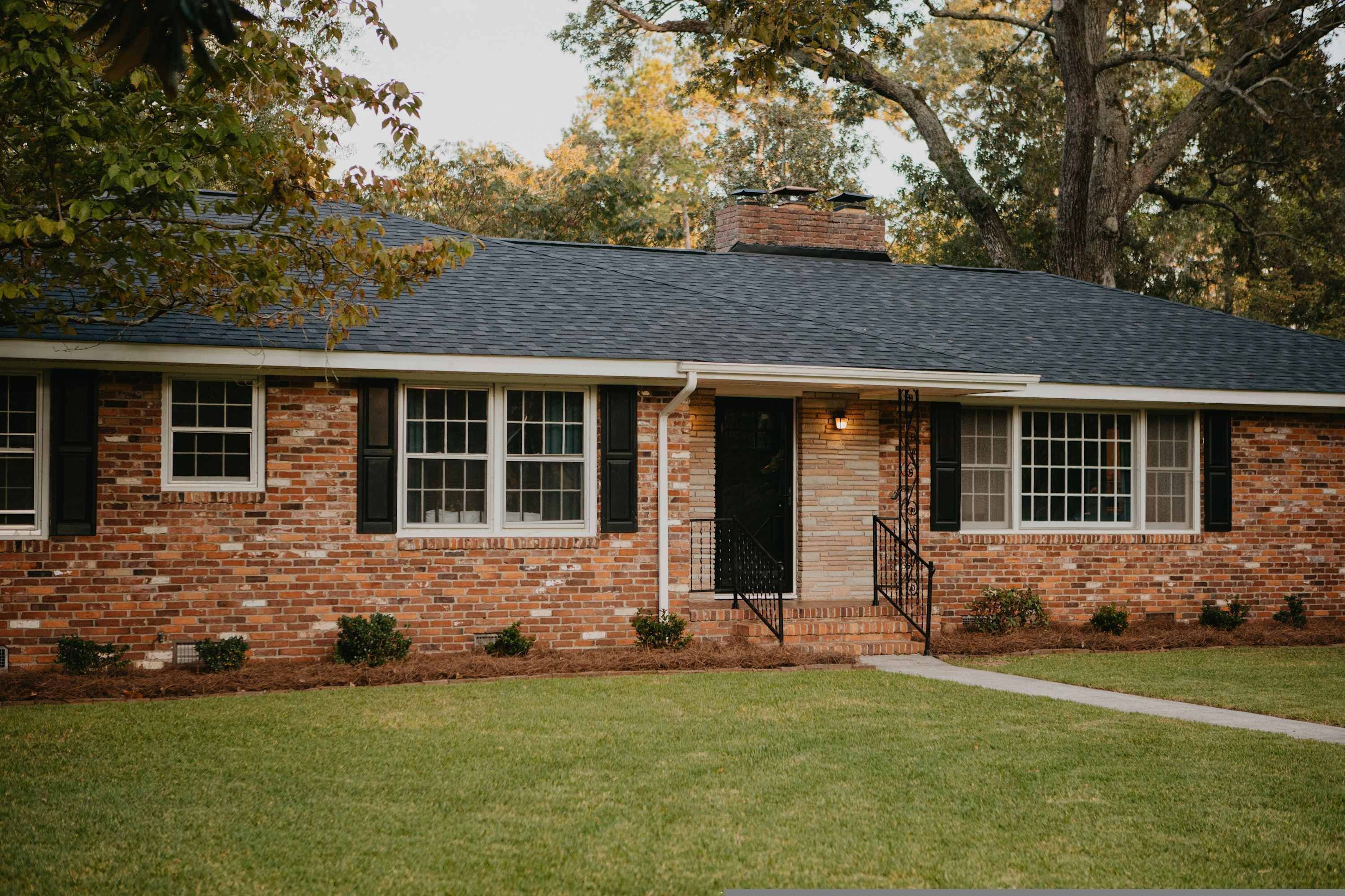 A single-story brick house with a gabled roof, a front porch with a staircase, and a small front yard.