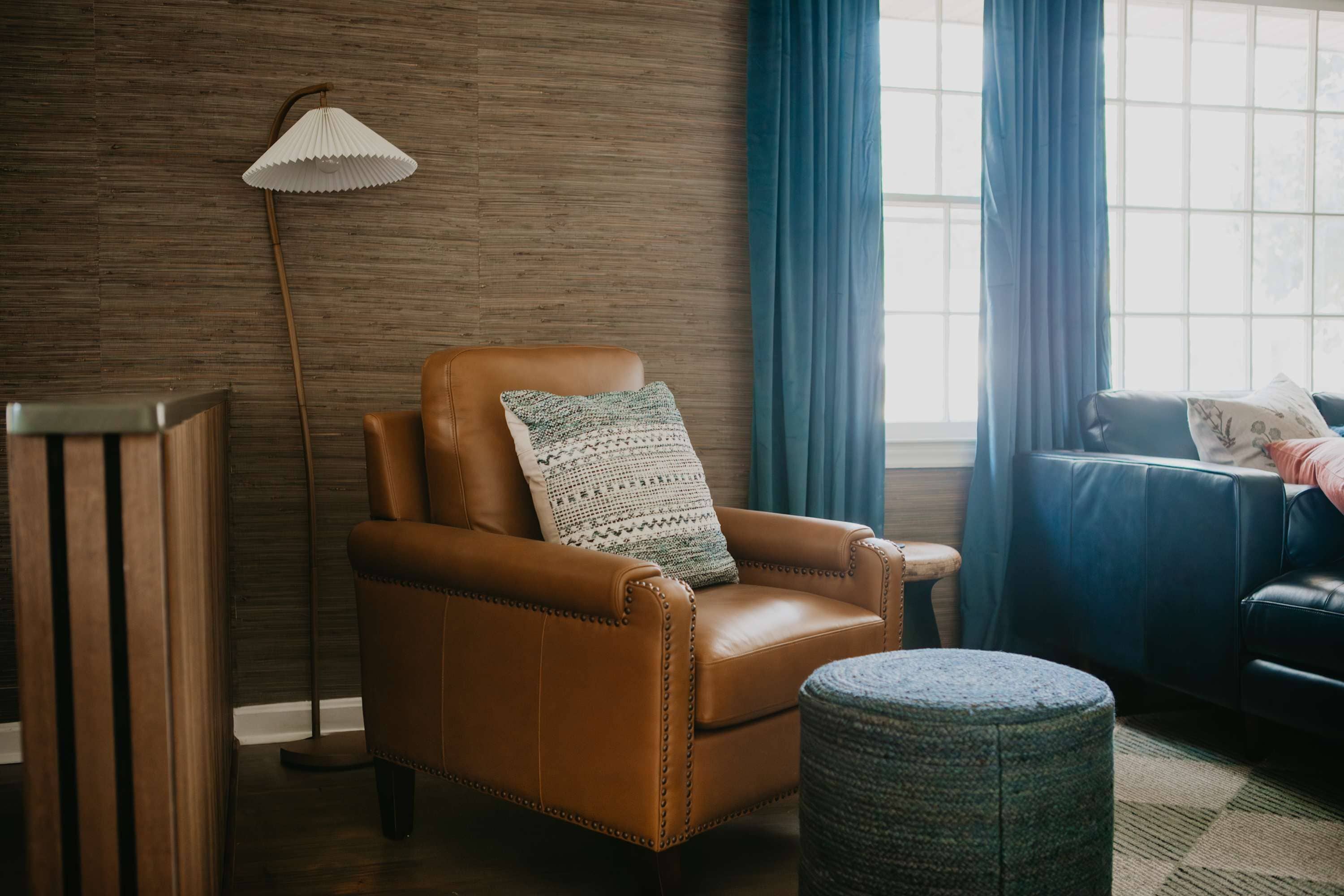 A room with a leather armchair, a floor lamp, a small ottoman, and a window with curtains.