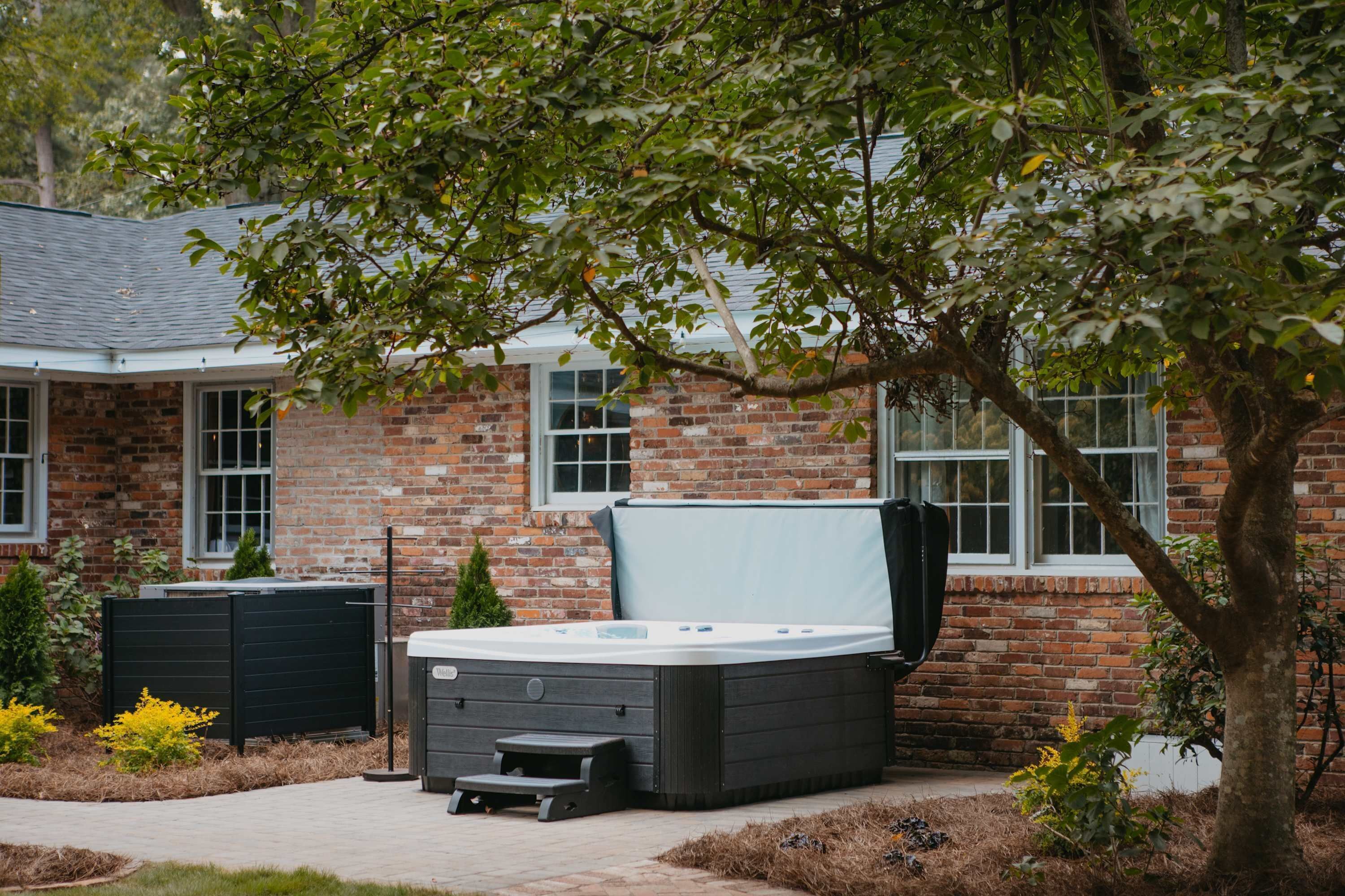 A hot tub in front of a brick house with a tree providing shade.