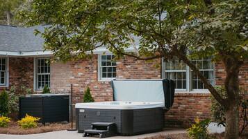 A hot tub in front of a brick house with a tree providing shade.