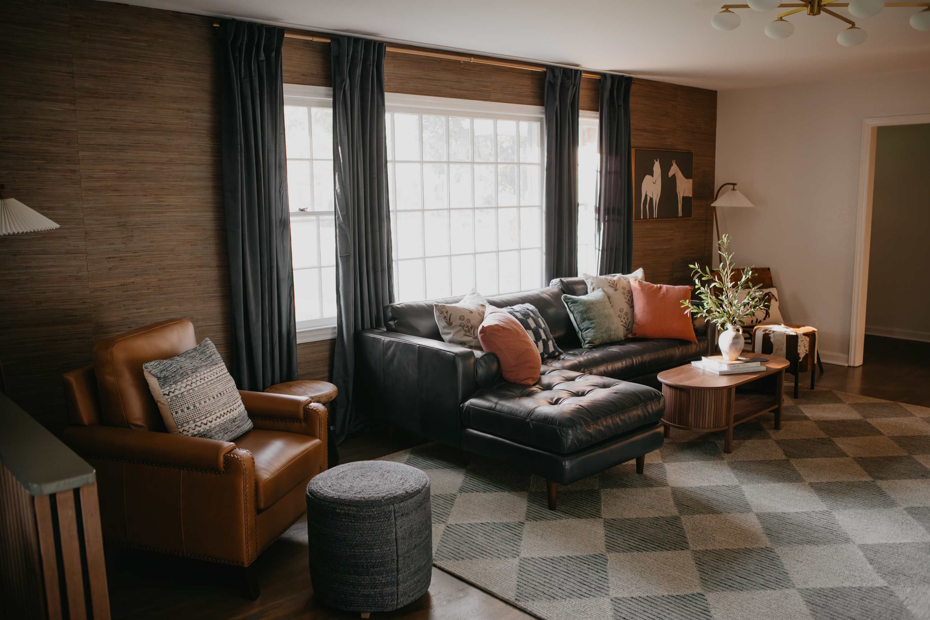 A living room with a leather sofa, a brown leather armchair, a round ottoman, and a coffee table with a book and a vase.