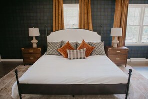 A neatly made bed with a white duvet, flanked by two nightstands with lamps, in a room with dark patterned walls and large windows with curtains. - Briarwood Cottage Hot Tub Putt Putt Games (Aiken)
