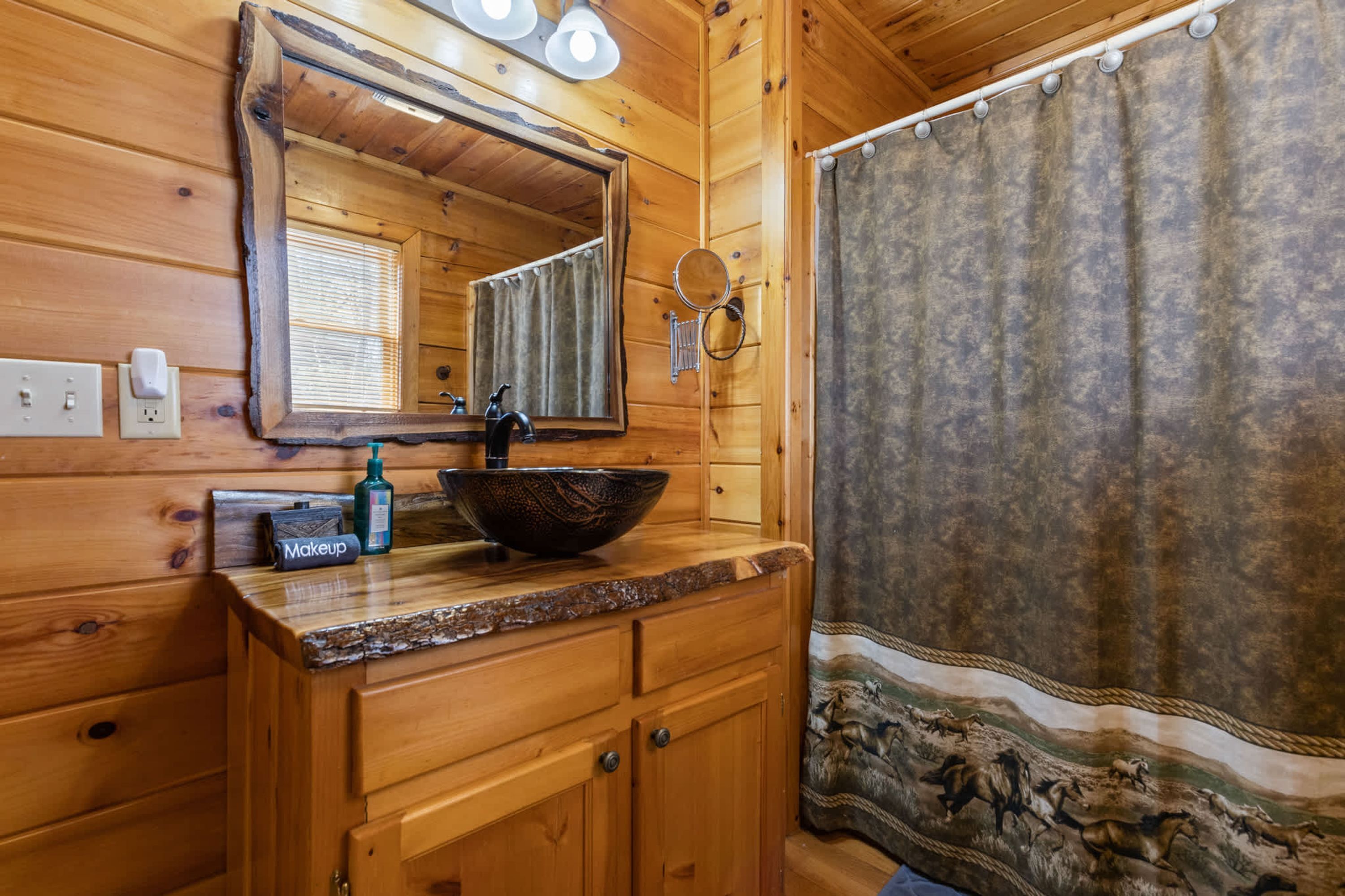 A wooden cabin bathroom with a wooden vanity, a large mirror, a vessel sink, and a shower curtain with a landscape design.