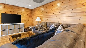 Interior acolhedor de uma cabana, com televisão de tela plana, um sofá, uma mesa de centro e uma luminária.