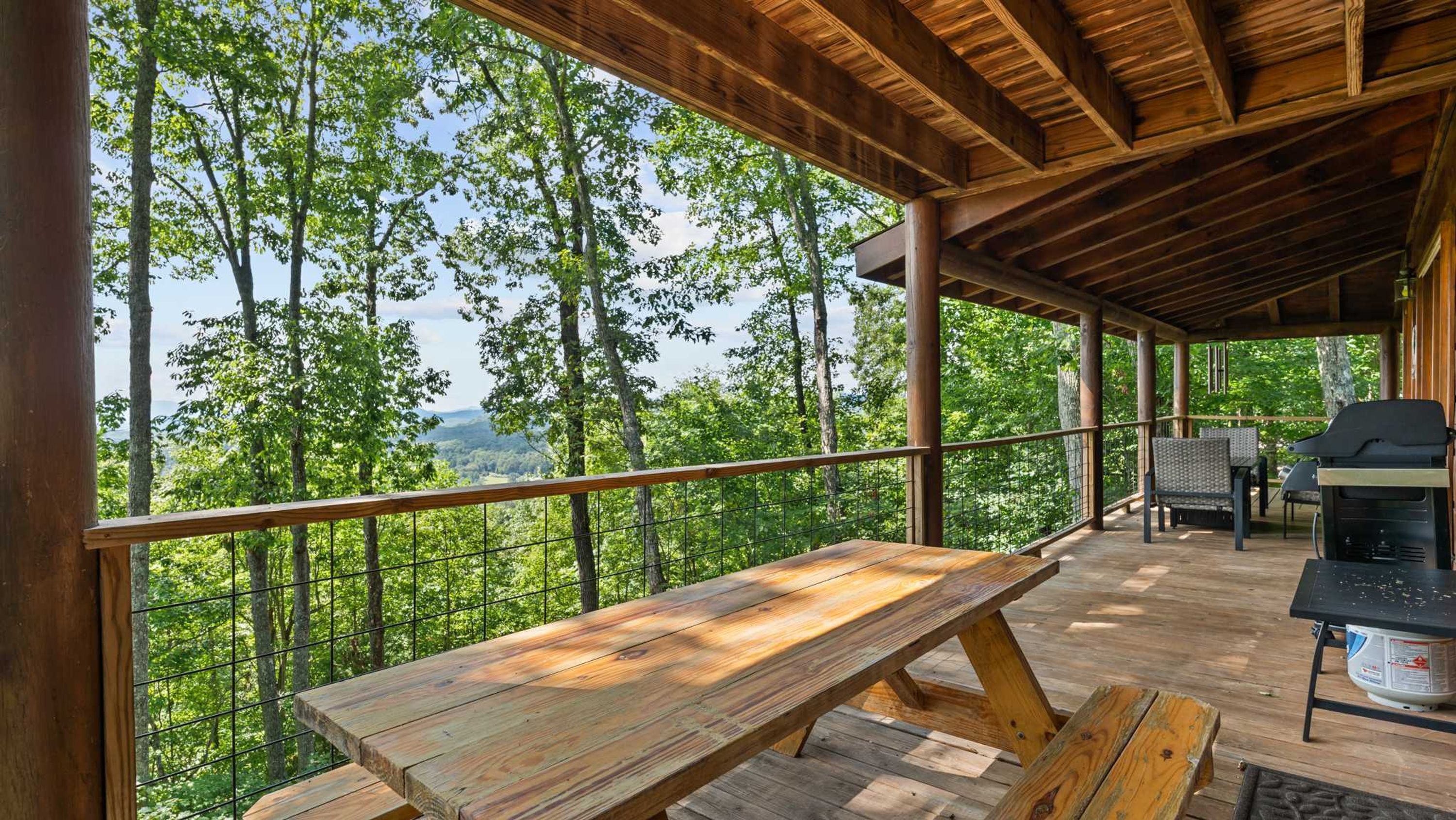 An outdoor wooden deck with a picnic table and grill, surrounded by trees and a view of the landscape.