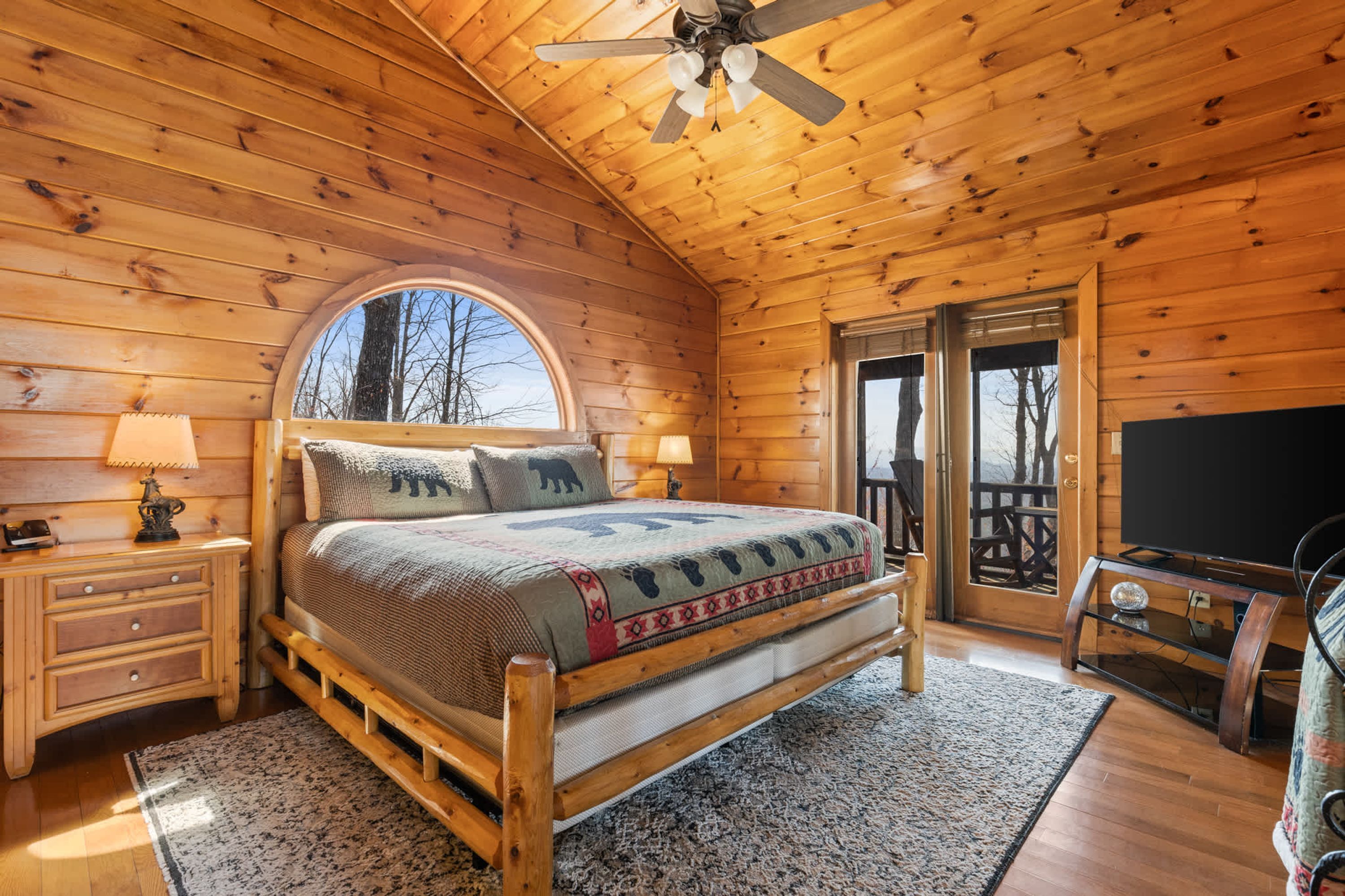 A wooden cabin bedroom with a bed, nightstands, a ceiling fan, and a large window.