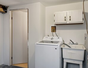 Un cuarto de lavandería con lavadora y secadora de carga frontal, un gabinete blanco sobre la secadora y una puerta que da a otra habitación.