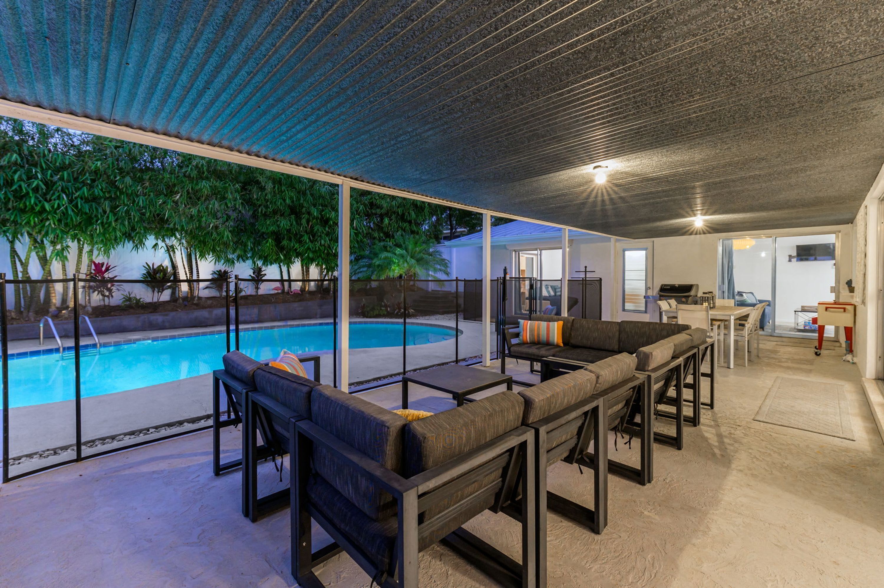 An outdoor pool area with a covered seating area and a kitchen visible through the glass doors.