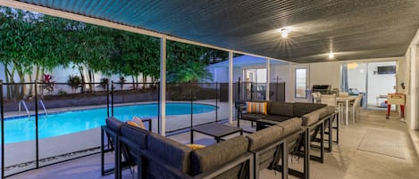 Zona de piscina exterior con un área de descanso cubierta y una cocina visible a través de puertas de cristal.