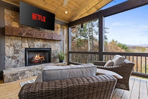Una chimenea de piedra con fuego, un cartel de Netflix encima, muebles de mimbre y vistas a árboles y montañas.