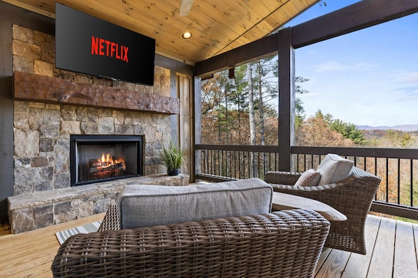 Una chimenea de piedra con fuego, un cartel de Netflix encima, muebles de mimbre y vistas a árboles y montañas.