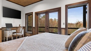 A bedroom with a bed, a desk with a chair, a TV mounted on the wall, and large windows with a view of trees. - 2-level Deck - Stunning Views - Hot Tub - Fire Pit (Ellijay)