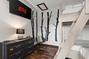 Un dormitorio con una cama individual superpuesta, un televisor montado en la pared, un aparador con cajones, una silla y una pared con siluetas de árboles.