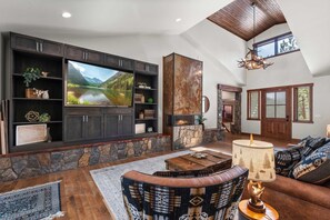 A spacious living room with a large flat-screen TV, stone fireplace, and a high ceiling with wooden beams. - 34 Acre Escape Games Hiking Pool Table Bicycles (Black Hawk)