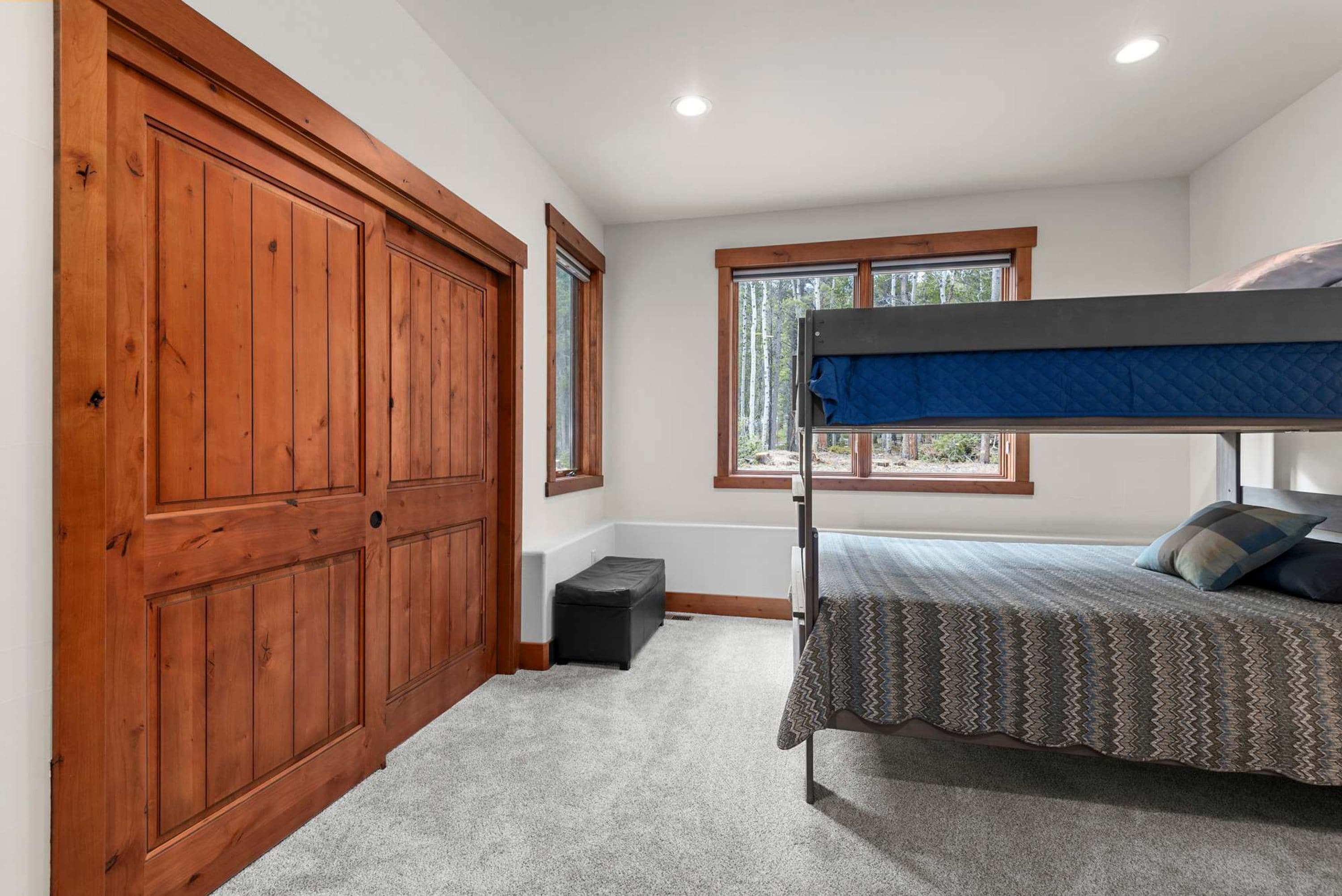 A bedroom with a bunk bed, a wooden wardrobe, a window with a view of trees, and a carpeted floor.