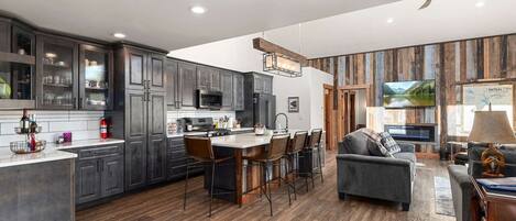 A modern kitchen with dark cabinets, a central island with bar stools, and a dining area with a sofa and TV.