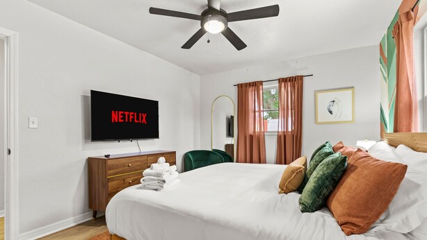 A hotel room with a bed, a television displaying 'NETFLIX,' a wooden dresser, a green chair, and a window with curtains. - Resort Style Backyard Pickleball Pool Putt Putt (Seminole)