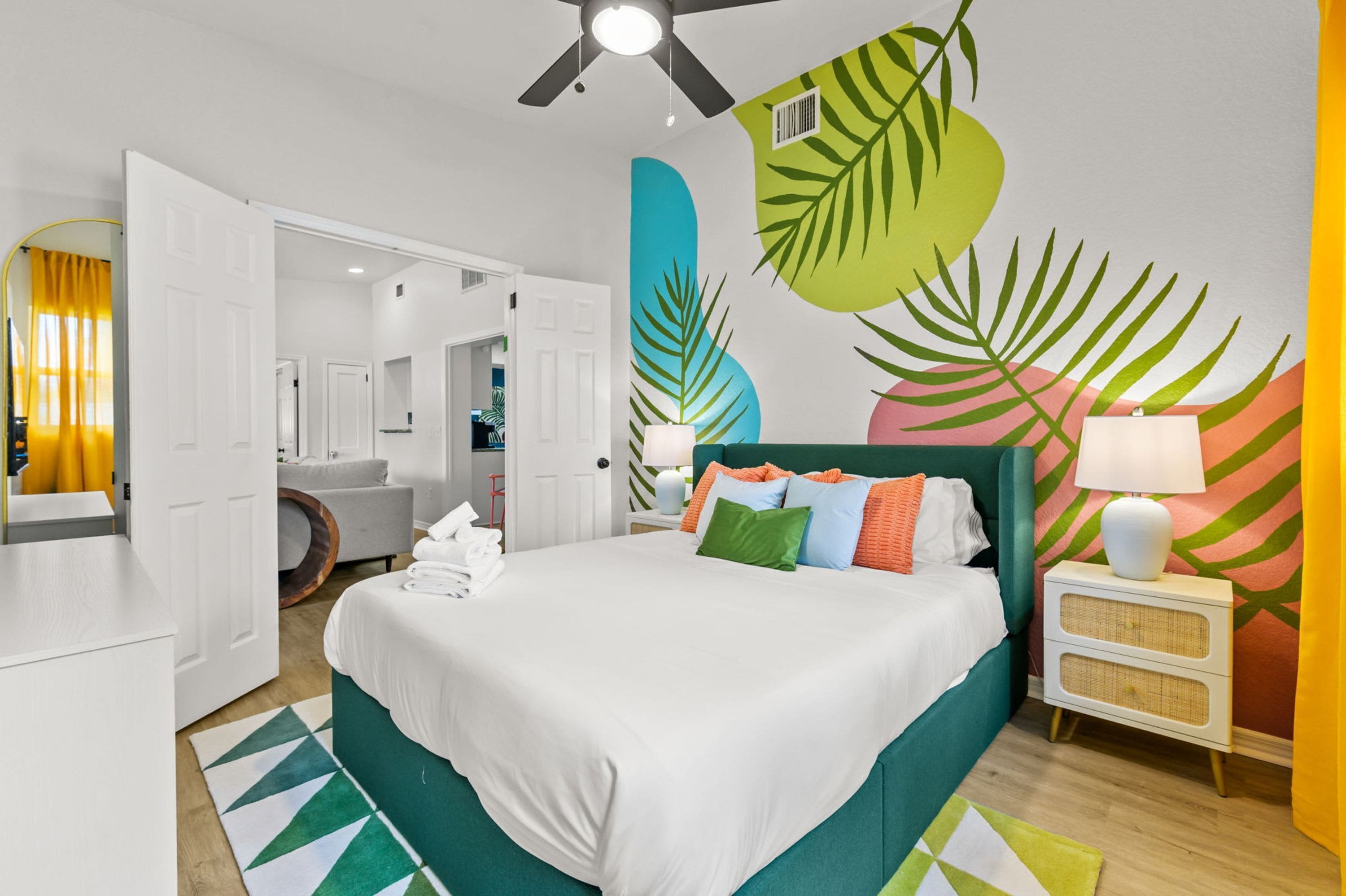 Ein Hotelzimmer mit einem Bett, einem Nachttisch, einer Lampe, einem Deckenventilator und einer Wand mit tropischen Pflanzenmotiven.