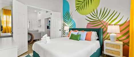 Une chambre d’hôtel comprenant un lit, une table de chevet, une lampe, un ventilateur de plafond et un mur décoré de motifs représentant des feuilles tropicales.