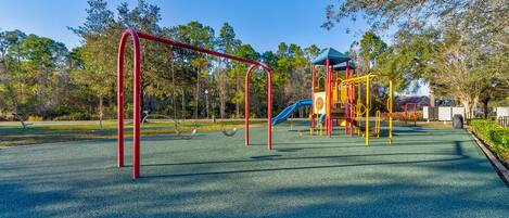 A playground with swings and a slide, surrounded by trees and a clear sky.