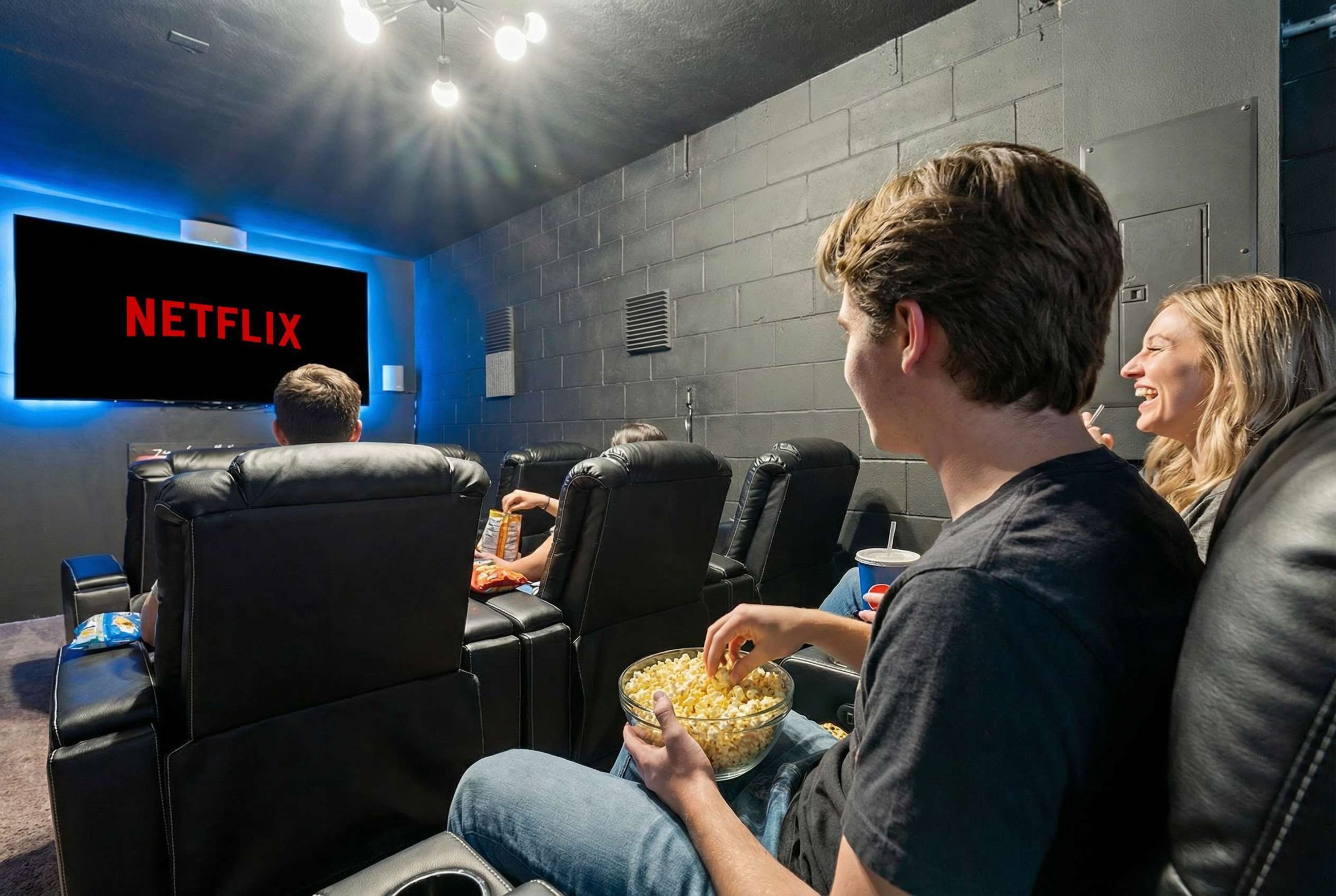 A movie theater with black leather seats, a large screen displaying 'NETFLIX', and a person holding a bowl of popcorn.