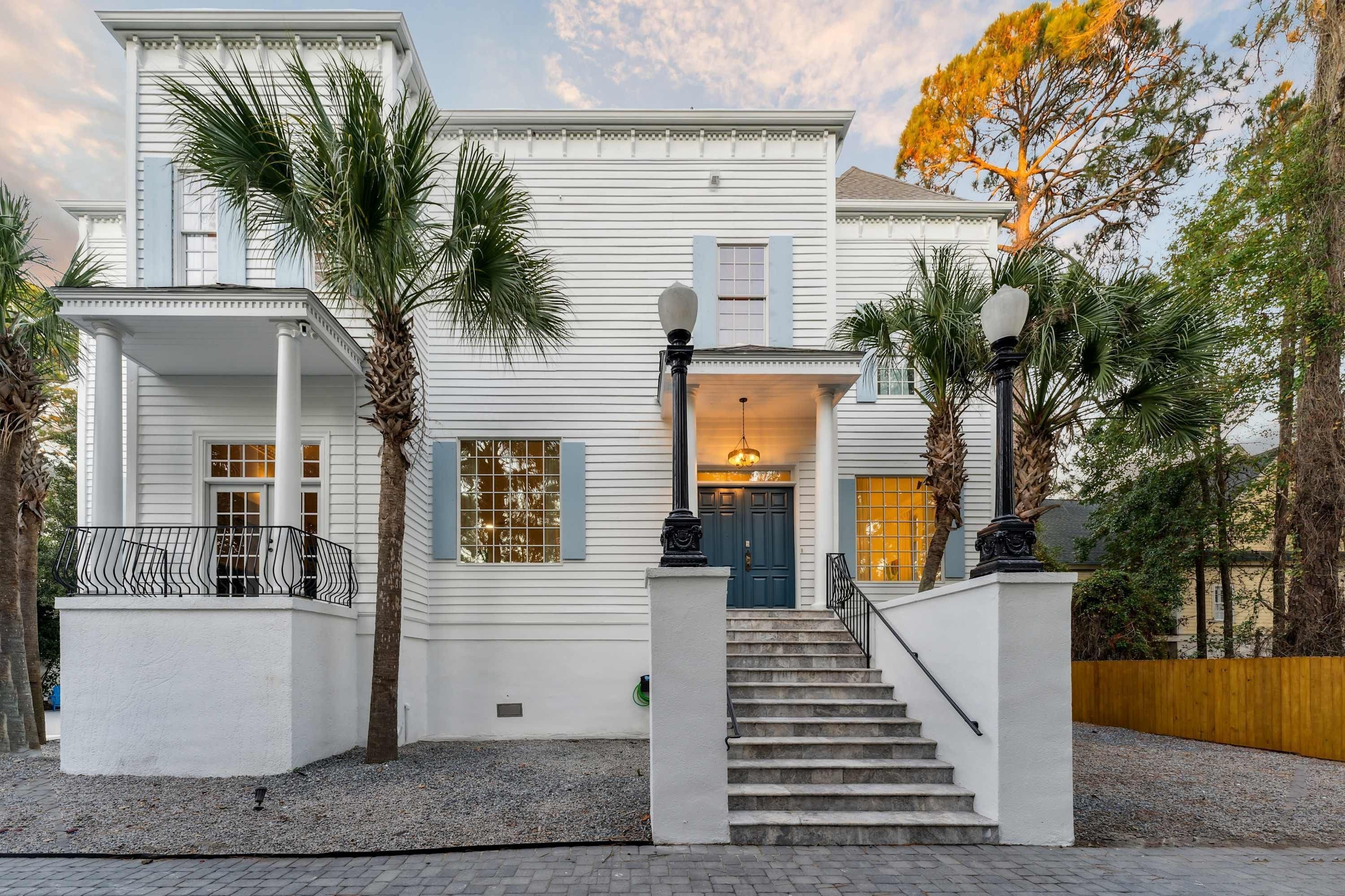 A two-story white house with a front porch, a staircase, and palm trees.