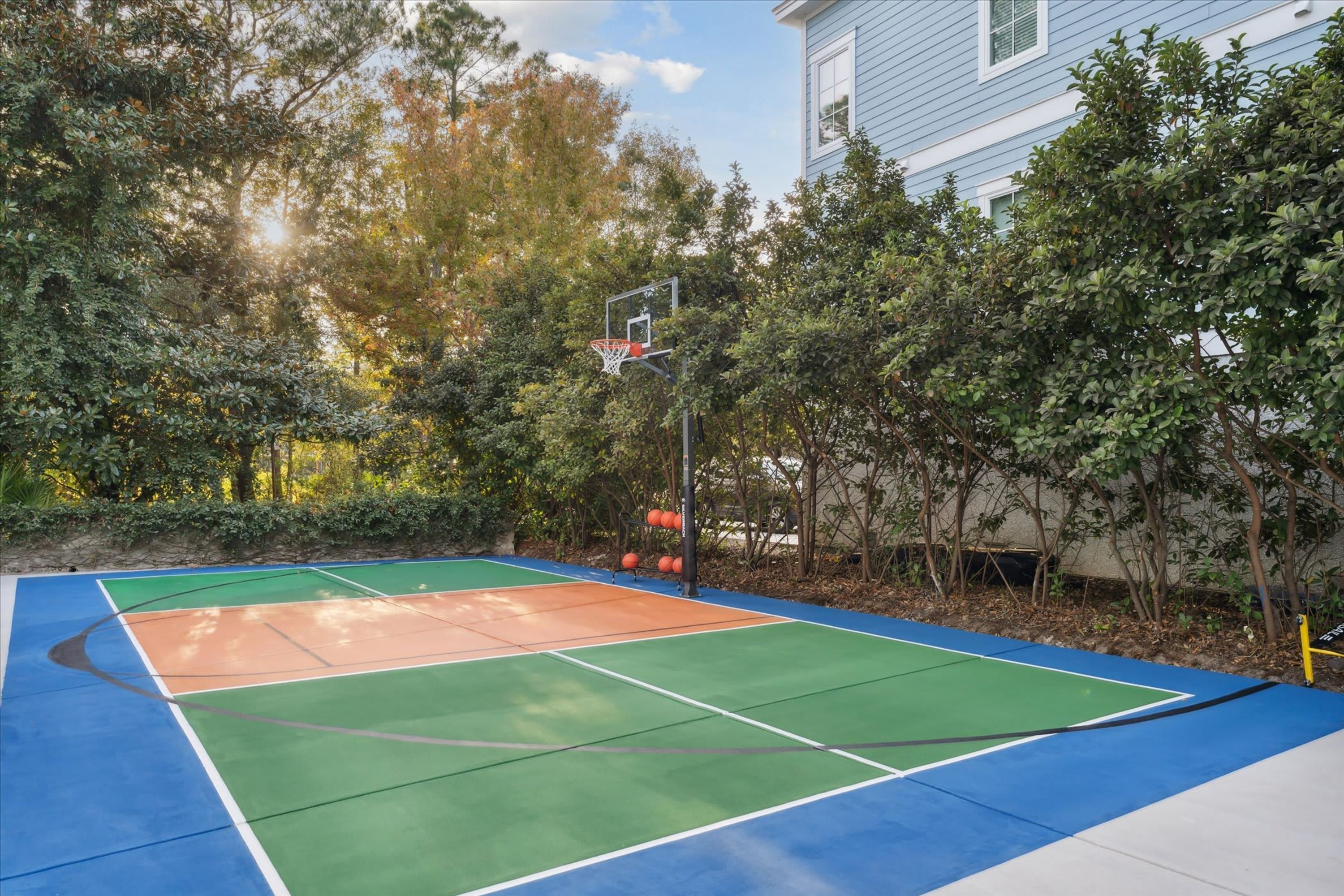 Een basketbalveld buiten, met een speeloppervlak in blauw, groen en oranje, omringd door bomen en een gebouw.
