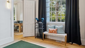 A room with a wooden floor, a bench with cushions, a window with curtains, and a door leading to another room. - Burkes Beach Reserve Golf Sim Pickleball Sleeps 24 (Hilton Head Island)