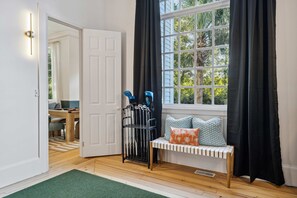 A room with a wooden floor, a bench with cushions, a window with curtains, and a door leading to another room. - Burkes Beach Reserve Golf Sim Pickleball Sleeps 24 (Hilton Head Island)