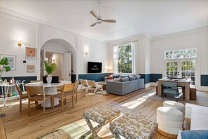 A spacious living room with a large grey sofa, a dining area with a white table and chairs, and a ceiling fan. - Burkes Beach Reserve Golf Sim Pickleball Sleeps 24 (Hilton Head Island)
