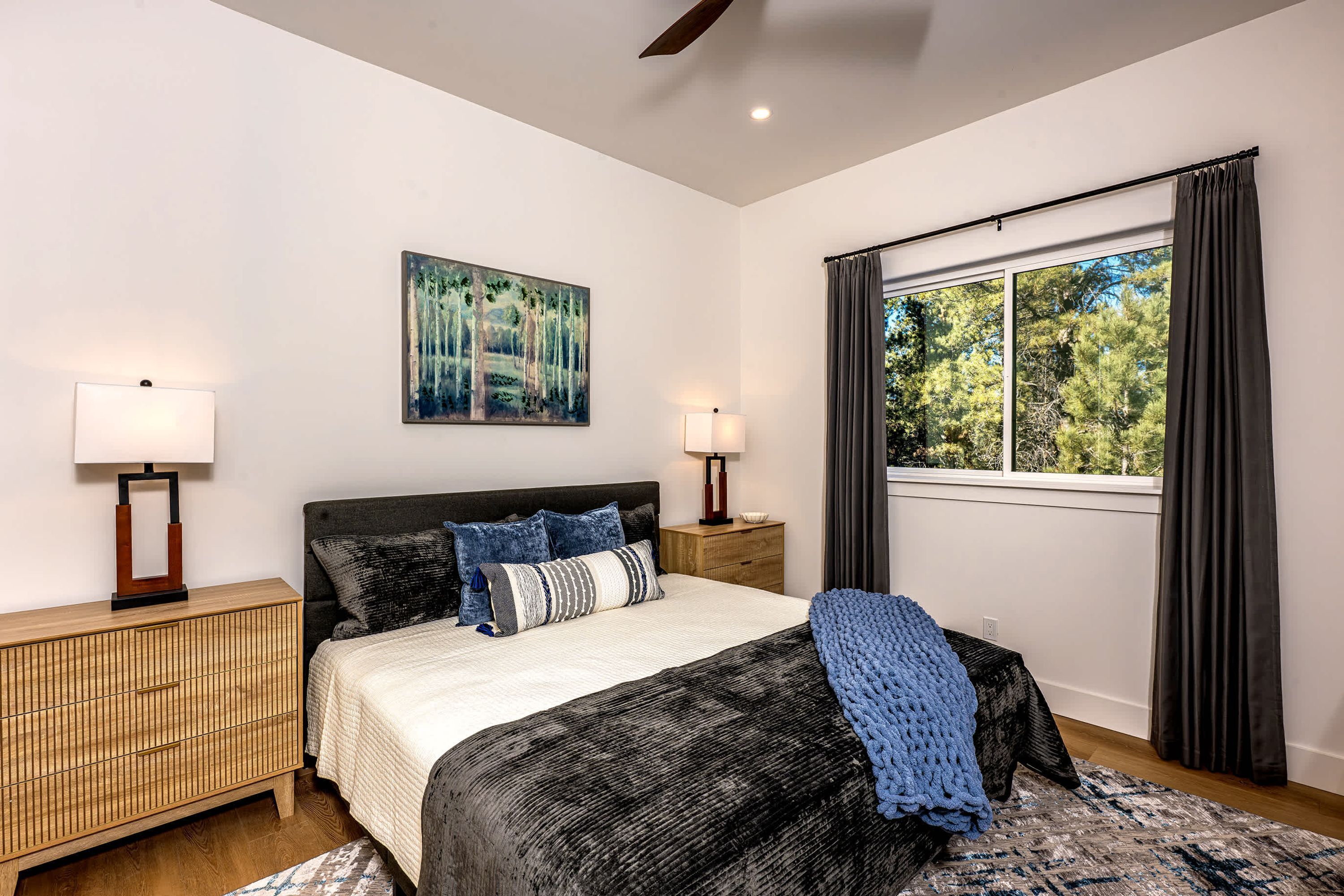 A bedroom with a bed, bedside lamps, a painting, a window with curtains, and a view of trees.