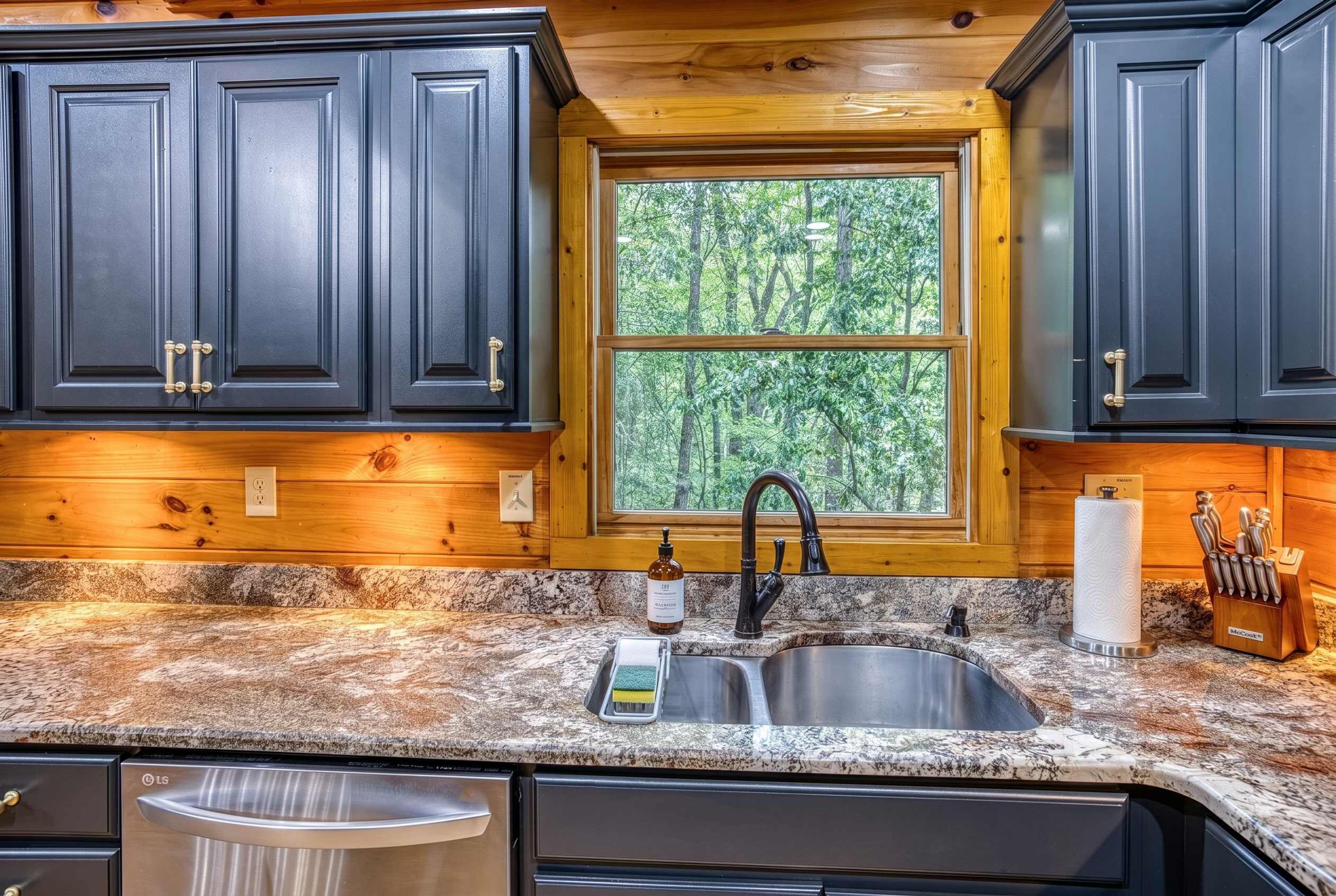 A kitchen with dark cabinets, a granite countertop, a stainless steel sink, and a window with a view of trees.